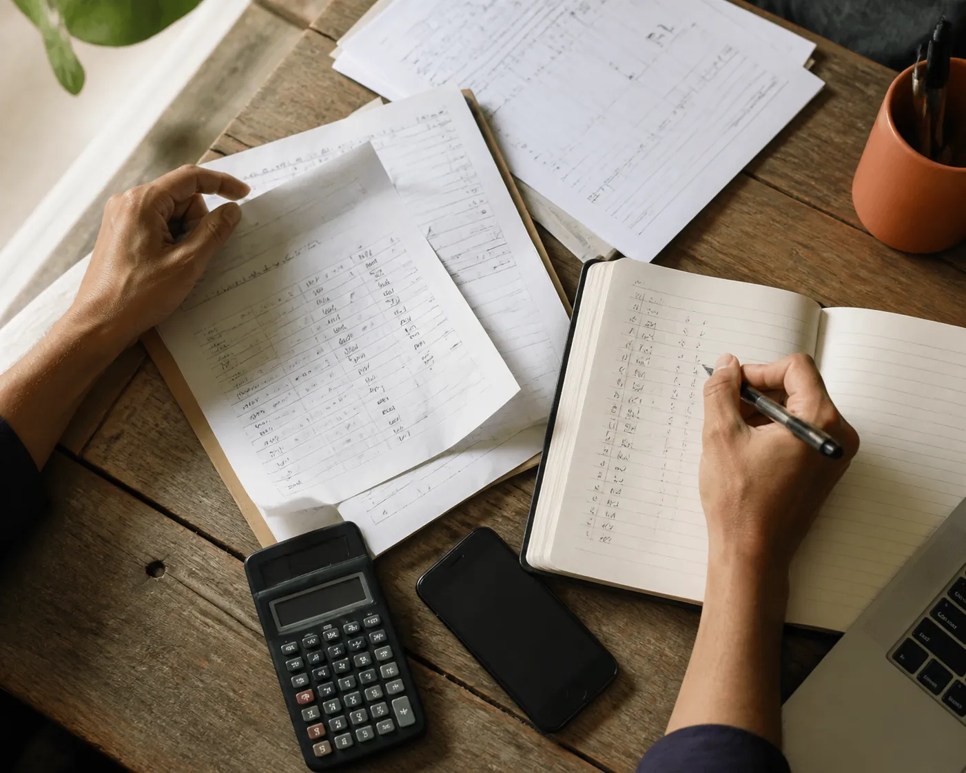 Manos revisando hojas de pedidos y anotando prioridades en una libreta, junto a una calculadora y un teléfono sobre una mesa de trabajo.
